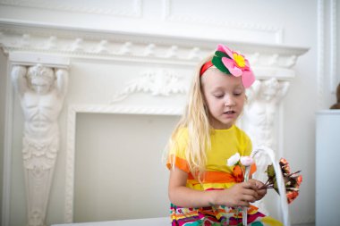 Güzel kız giyiyor parlak sarı elbise, pembe süs, aksesuar. Anneler günü tebrik ederim. Eğlenceli ve neşeli bir çocuk. Beyaz sepet çiçekli. Gülümse ve iyi bir ruh hali için pozitif sürpriz