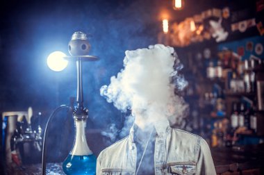 Adam barda nargile içiyor ve yüzünün önünde duman üflediyor.