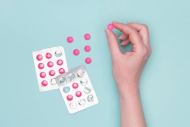 Top view of female hand holding pink prescription pill next to blister packs over pastel blue background. Sick patient taking medication. clipart