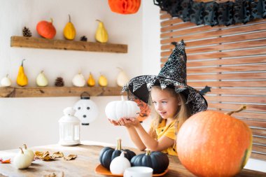 Sevimli küçük kız Halloween tema içinde bir masanın arkasında oturan cadı kostüm odası, boyalı kabaklar ve gülümseyerek elini tutarak dekore edilmiş. Cadılar Bayramı tatil kavramı.
