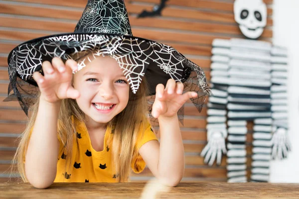 ハロウィーンのテーマのテーブルの後ろに座っている魔女帽子をかぶってかわいい女の子は 怖い顔をしてカメラ目線のリビング ルームを装飾されています ハロウィーン パーティー コンセプト — ストック写真