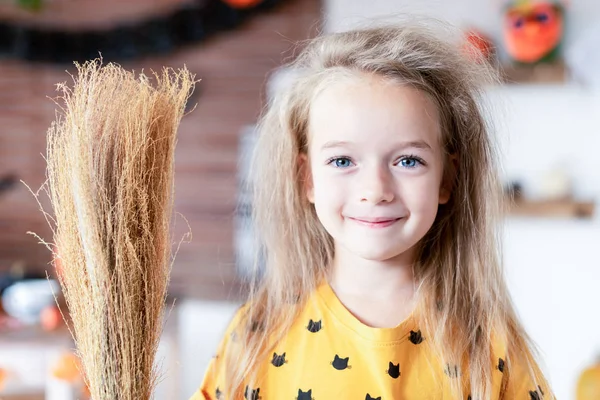 Cute little girl with messy hair, dressed up as a witch and holding a broom is standing in Halloween decorated living room, looking at camera smiling. Halloween party concept.