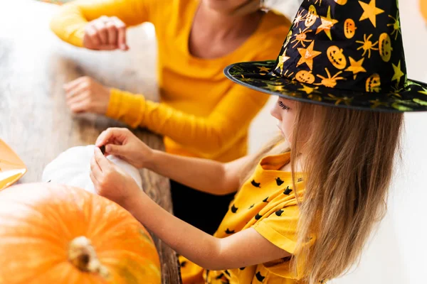 身に着けている白のハロウィンかぼちゃを飾る彼女の母親と一緒にテーブルの後ろに魔女帽子座っているかわいい女の子 ライフ スタイル家族率直なハロウィーンのテーマ背景 — ストック写真