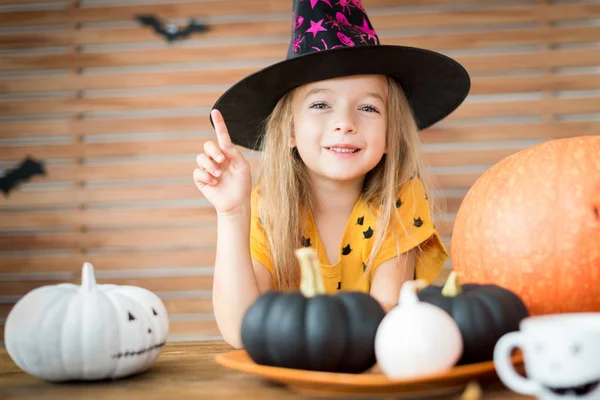魔女の帽子をかぶってかわいい女の子は ハロウィンのカボチャとテーブルの後ろに座っています 笑みを浮かべて 彼女の指が上向き カメラ目線のハロウィーンの衣装の女の子 — ストック写真