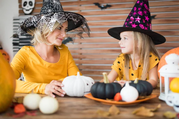 かわいい女の子と彼女の母親 両方魔女衣装 ハロウィーンのテーマでテーブルの後ろに座っているのお互いを見て 笑みを浮かべての部屋を飾った 家族のハロウィーン ライフ スタイル コンセプト — ストック写真