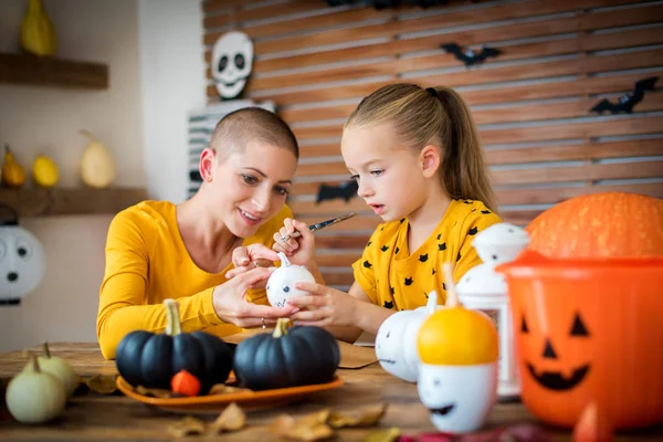 Cute young girl sitting at a table, decorating little white pumpkins with her mother, a cancer patient. DIY Halloween holiday and family lifestyle background.