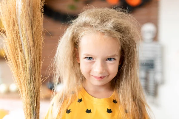 Cute little girl with messy hair, dressed up as a witch and holding a broom is standing in Halloween decorated living room, looking at camera smiling. Halloween party concept.