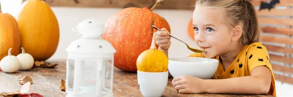 Child eating pumpkin soup in Halloween decorated dinning room. Autumn season food lifestyle banner.