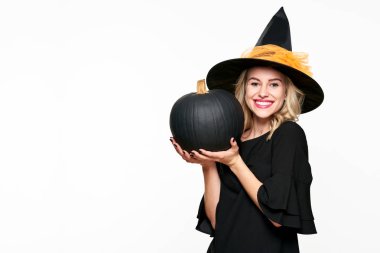 Halloween cadı büyük siyah kabak tutan gülümseyerek muhteşem. Cadı şapka giymiş güzel bir genç kadının portresi. Cadılar Bayramı kavram beyaz arka plan üzerinde.