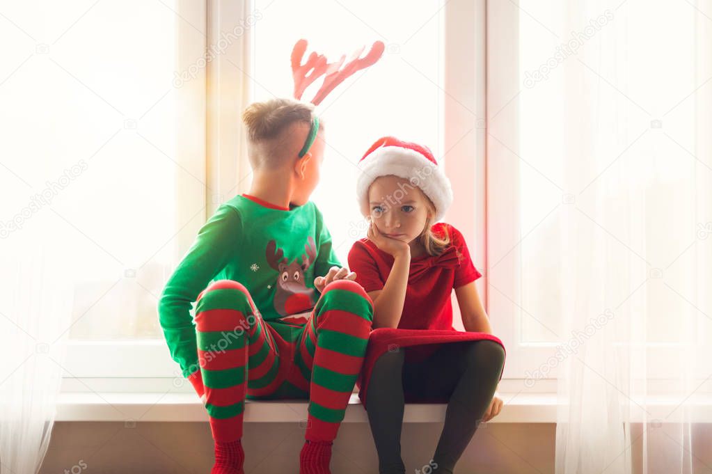 Brother and sister sitting restless on window sill at christmas time, looking out the window, anxiously waiting for Santa Claus to come. Christmas family time lifestyle.