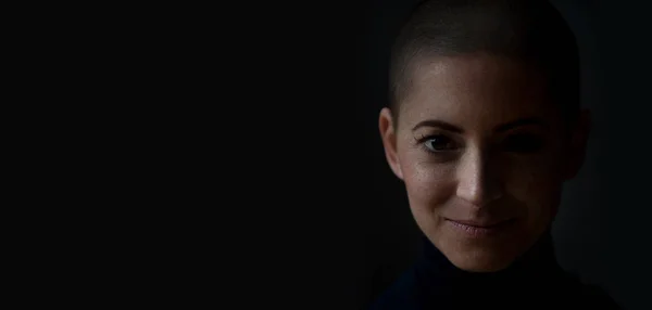 Portrait of a beautiful young courageous smiling female cancer patient, with shaved head. Gorgeous woman, a cancer patient, portrait on dark background with copy space.