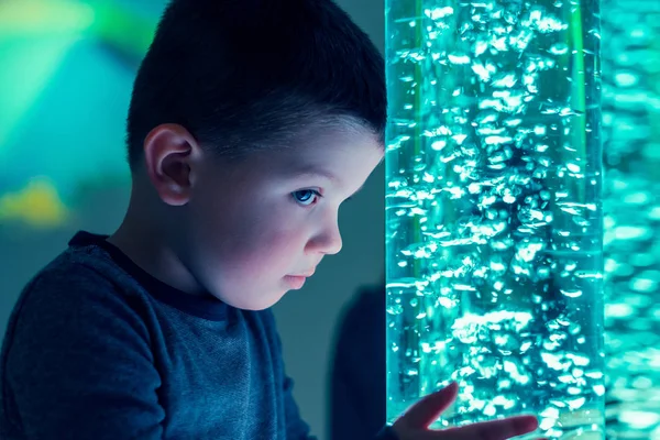 Child in therapy sensory stimulating room, snoezelen. Child interacting with colored lights bubble tube lamp during therapy session.