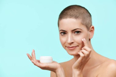 Mid 30s gülümseyen yüzünde nemlendirici krem uygulamak kadın. Kafası tıraşlı ve sağlıklı cilt üzerinde pastel mavi arka plan izole çekici beyaz kadın fotoğrafı. Güzellik ve cilt bakımı kavramı.