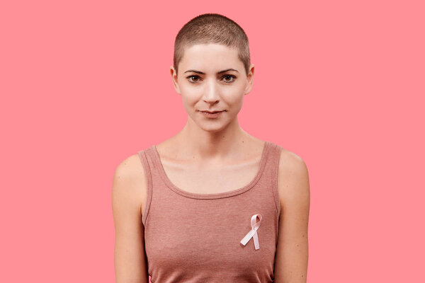 Улыбающаяся женщина за 30, пережившая рак, носящая розовую ленту для информирования о раке молочной железы, изолированная на живом коралловом фоне. Поддержка, солидарность, скрининг и профилактика
.