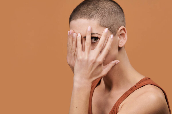 Beautiful young woman with shaved head covering her face with both hands and peeking through fingers. Woman with frightened expression isolated over brown background.