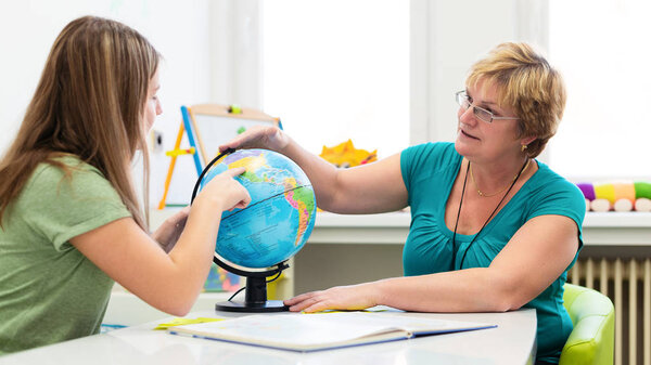 Mature female therapist working with a teenage girl with learning difficulties. Private geography tutoring session. Teacher with teenage student in class pointing to countries on a globe.