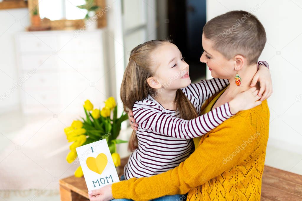 Adorable young girl giving her mom, young cancer patient, homemade I LOVE MOM greeting card. Family celebration concept. Happy Mother's Day or Birthday Background. 