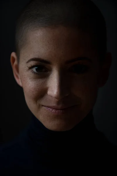 Portrait of a beautiful young courageous smiling female cancer patient, with shaved head. Gorgeous woman, a cancer patient, portrait on dark background with copy space.