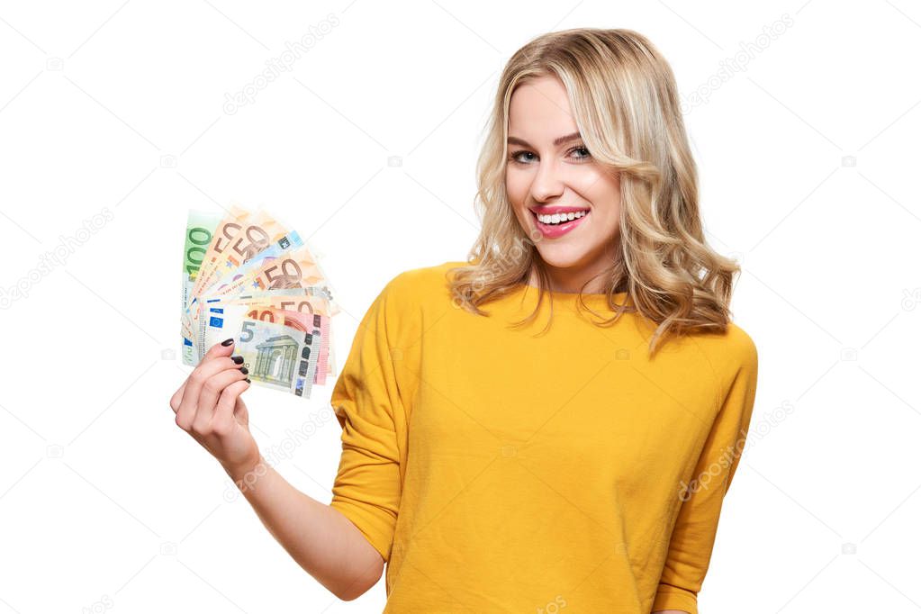 Young pretty woman in yellow sweater holding bunch of Euro banknotes, looking at camera and smiling, isolated on white background. 