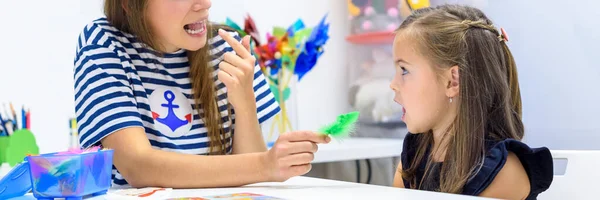Çocuk konuşma terapisi konsepti. Anaokulu öğrencisi bir kadın konuşma terapistiyle doğru telaffuz pratiği yapıyor. Web pankartı.