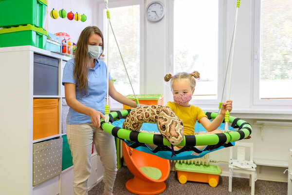 Toddler girl in child occupational therapy session doing playful exercises with her therapist during Covid - 19 pandemic, both wearing protective face masks.