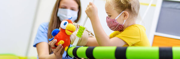 Toddler girl in child occupational therapy session doing playful exercises with her therapist during Covid - 19 pandemic, both wearing protective face masks. Web banner.