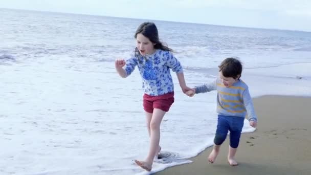 Niños felices jugando en la playa. Hermana y su hermano pequeño corren tomados de la mano — Vídeo de stock