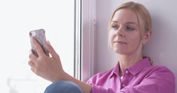 Menina loira bonita sentada em uma janela e smartphone em mãos — Vídeo de Stock