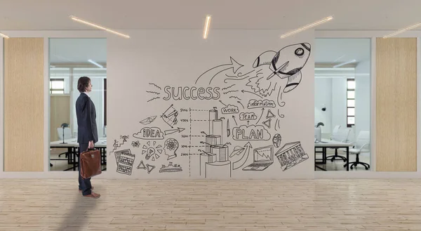 서류 가방을 가지고 있는 사업가가 현대식 사무실 의벽에 그려진 사업 도면을 보고 있다 — 스톡 사진