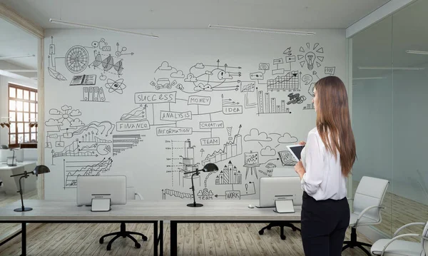 En affärskvinna står i moderna kontor med kreativ affärsstrategi skiss ritad på vit vägg — Stockfoto