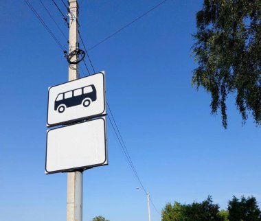 Bir otobüs durağı tabelası direğin üzerinde duruyor, toplu taşıma durağına işaret veriyor. Gökyüzü açık mavi, beyaz ve siyah işaretler için açık bir zemin sağlıyor..