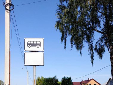 Bir otobüs durağı tabelası parlak mavi bir gökyüzünün altında bir elektrik direğinin yanında duruyor. Yakın ağaçlar ve evler gün boyunca sakin bir banliyö atmosferini resmediyor.