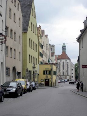 Regensburg 'da gezinirken renkli binalar ve bulutlu bir gökyüzü altında tarihi mekanlar sergileniyor. Yayalar canlı şehir atmosferinin keyfini çıkarıyorlar..