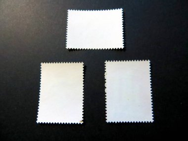 Üç adet eski posta pulu, boş ters yüzleri görünür bir şekilde sergileniyor, eşsiz dokularını ve kenarlarını vurguluyor, fitil meraklıları için ideal..