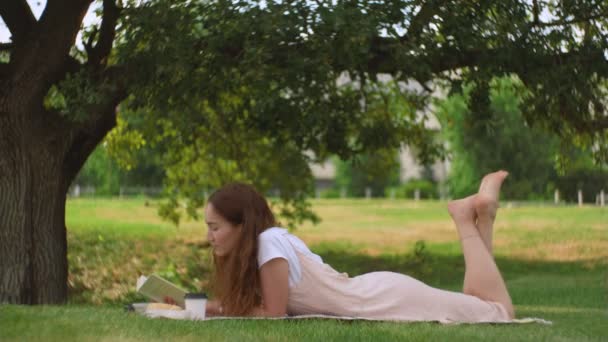 Genç bir kadın yalan söylüyor ve ağacın altında kitap okuyor. — Stok video