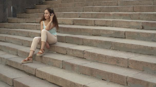 Attraktiv brunett flicka lyssnar musik och sitter på trappan utanför — Stockvideo