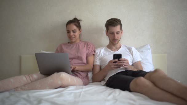 Glada par med hjälp av en dator och mobiltelefon ligger på sin säng hemma — Stockvideo