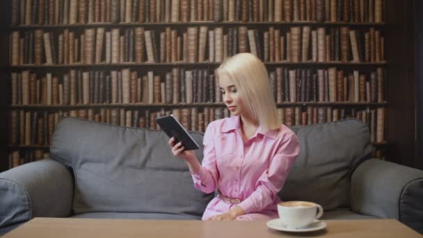 Blond vrouw in roze shirt is ebook lezen in cafe — Stockvideo