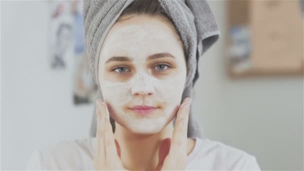 Красивая женщина с полотенцем на голове наносит косметическую маску и смотрит в камеру — стоковое видео