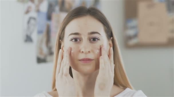 Bella giovane donna che applica la crema sul viso e guarda la fotocamera — Video Stock