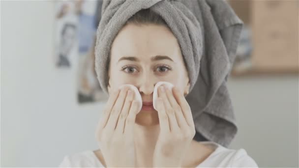 Mulher jovem com toalha na cabeça rosto de limpeza com almofadas de algodão e olhar para a câmera — Vídeo de Stock