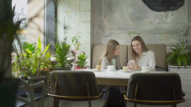 İki genç kadın telefona bakıyor ve kafede gülümsüyor.