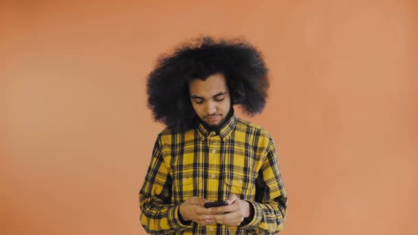 オレンジの背景に携帯電話を使っている若いハンサムなアフリカ系アメリカ人男性。感情の概念 — ストック動画