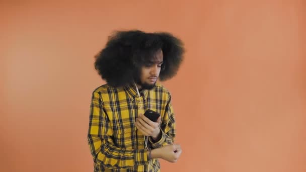 Uomo afroamericano positivo che usa il telefono per ascoltare musica nelle cuffie su sfondo arancione. Concetto di emozioni — Video Stock