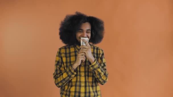 Feliz joven afroamericano hombre sosteniendo el dinero en sus manos y mirando a la cámara en el fondo naranja . — Vídeos de Stock