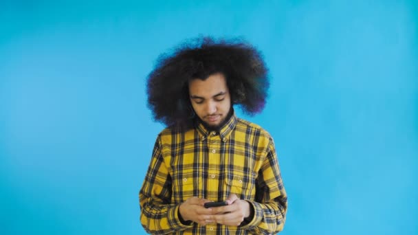 Joven hombre afroamericano guapo usando el teléfono sobre fondo azul. Concepto de emociones — Vídeos de Stock