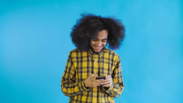 Ritratto di uomo afroamericano con capelli ricci che chatta su smartphone su sfondo blu. Concetto di emozioni — Video Stock
