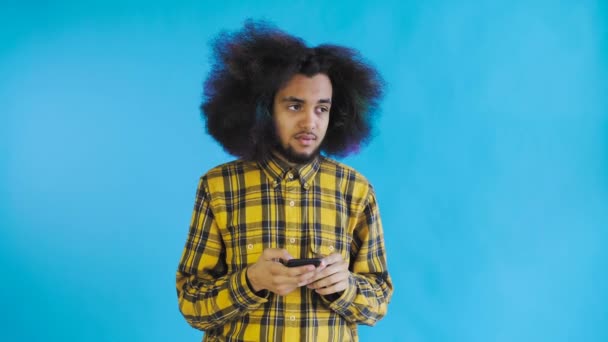 Hombre afroamericano con el pelo rizado está pensando antes de enviar un mensaje sobre fondo azul. Concepto de emociones — Vídeos de Stock