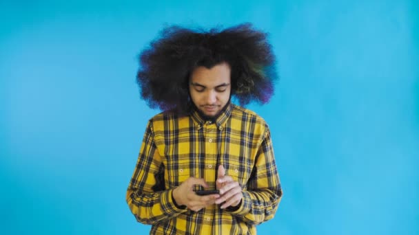 Joven afroamericano frustrado sosteniendo el teléfono móvil roto sobre fondo azul. Concepto de emociones — Vídeos de Stock