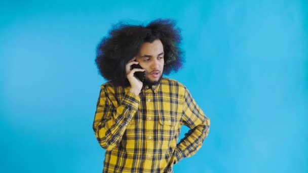 Feliz joven y atractivo afroamericano hablando por teléfono móvil sobre fondo azul. Concepto de emociones — Vídeos de Stock